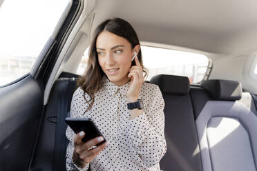 Female professional talking through in-ear headphones while holding smart phone in car - FMOF01388