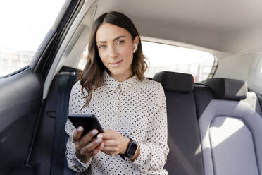 Geschäftsfrau mit In-Ear-Kopfhörern hält Smartphone im Auto - FMOF01389