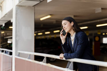 Weibliche Unternehmerin spricht mit ihrem Smartphone, während sie sich auf einem Parkplatz an ein Geländer lehnt - FMOF01405