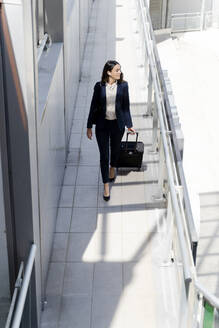 Weibliche Unternehmerin schaut weg, während sie mit einem Koffer durch einen Gebäudeflur geht - FMOF01409