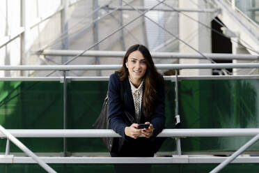 Businesswoman with smart phone leaning on railing in building - FMOF01410