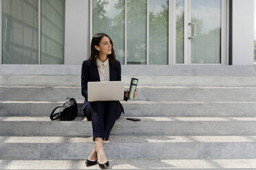 Businesswoman with laptop looking away holding travel mug while sitting on staircase - FMOF01413