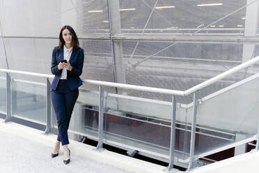 Geschäftsfrau mit Smartphone, die sich am Geländer eines Bürogebäudes anlehnt - FMOF01417