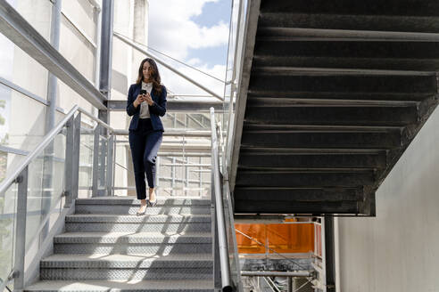 Geschäftsfrau, die ein Smartphone benutzt, während sie eine Treppe in einem Gebäude hinuntergeht - FMOF01427