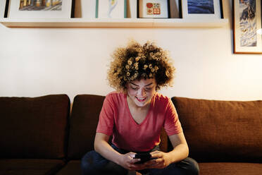 Smiling young with curly hair text messaging through smart phone in living room - ASGF00306
