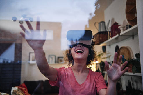 Fröhliche junge Frau, die einen Virtual-Reality-Simulator trägt, gesehen durch Glas zu Hause - ASGF00309