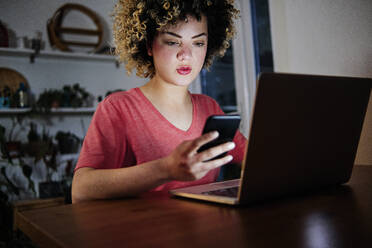 Beautiful young woman sitting with laptop using smart phone at home - ASGF00310