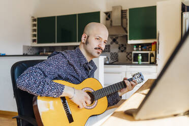Männlicher Gitarrist schaut auf den Computer, während er im Wohnzimmer Gitarre spielt - MEUF03018