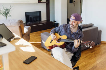 Lächelnder männlicher Gitarrist mit Mütze, der zu Hause Gitarre spielt - MEUF03019