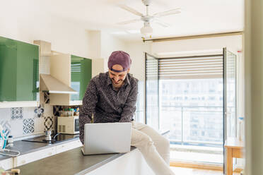 Smiling young man using laptop while sitting on kitchen island at home - MEUF03022