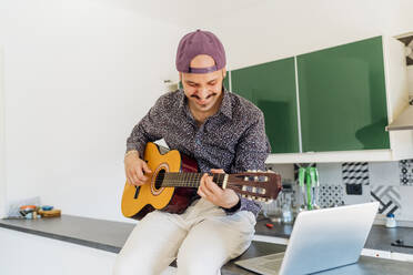 Smiling male composer playing guitar by laptop on kitchen island at home - MEUF03025