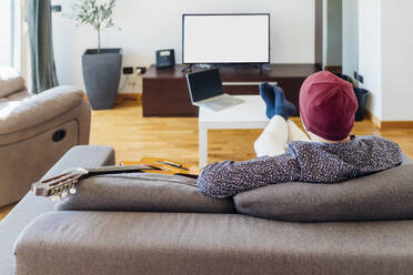 Männlicher Gitarrist beim Fernsehen auf dem Sofa zu Hause - MEUF03027