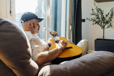 Male composer playing guitar while sitting on reclining chair at home - MEUF03029