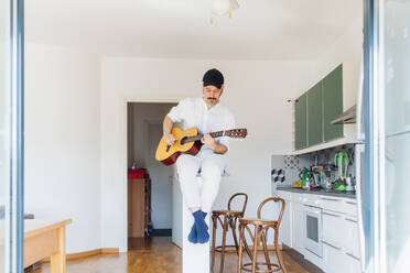 Male guitarist wearing cap playing guitar at kitchen - MEUF03031