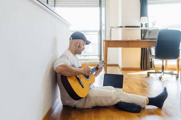 Männlicher Gitarrist spielt Gitarre am Laptop, während er zu Hause auf dem Boden sitzt - MEUF03033