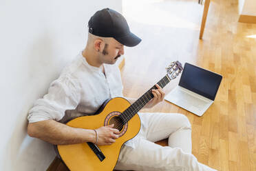 Man wearing cap playing guitar while sitting on floor at home - MEUF03034