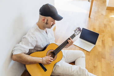 Mann mit Mütze spielt Gitarre, während er zu Hause auf dem Boden sitzt - MEUF03034