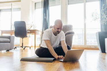 Junger Mann benutzt einen Laptop, während er zu Hause auf einer Übungsmatte sitzt - MEUF03048