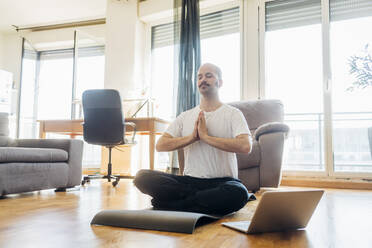 Man with hands clasped meditating while sitting on mat at home - MEUF03049