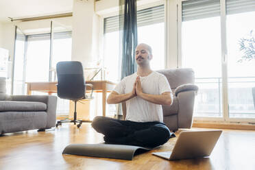 Mann mit verschränkten Händen meditiert auf einer Matte sitzend zu Hause - MEUF03049
