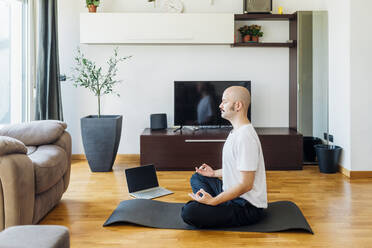 Bald man in lotus position sitting on exercise mat at home - MEUF03050