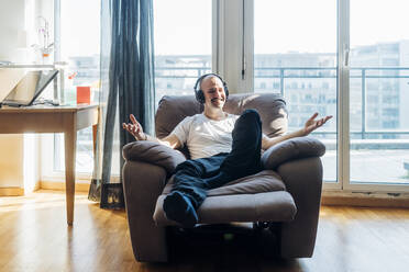 Lächelnder Mann mit Kopfhörern, der auf einem Lehnstuhl im Wohnzimmer sitzt und gestikuliert - MEUF03053