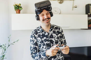 Glücklicher Mann mit Virtual-Reality-Headset, der zu Hause einen Game-Controller hält - MEUF03063