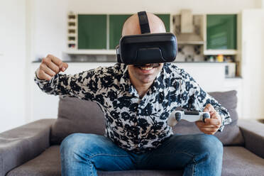 Lächelnder Mann, der ein Virtual-Reality-Headset trägt, während er auf dem Sofa zu Hause einen Game-Controller hält - MEUF03064