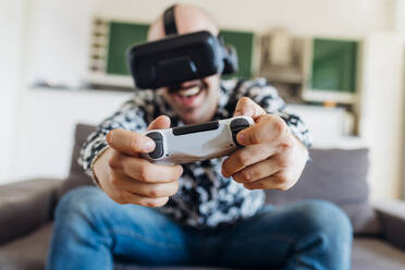 Happy man wearing virtual headset playing video game on sofa - MEUF03065