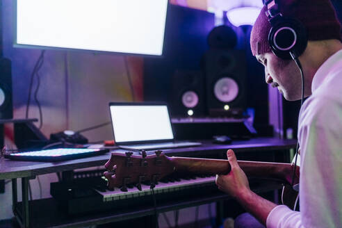 Man wearing headphones playing guitar at home studio - MEUF03099