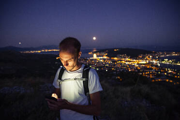 Man using mobile phone in front of town at night, Trogir, Croatia - MAMF01823