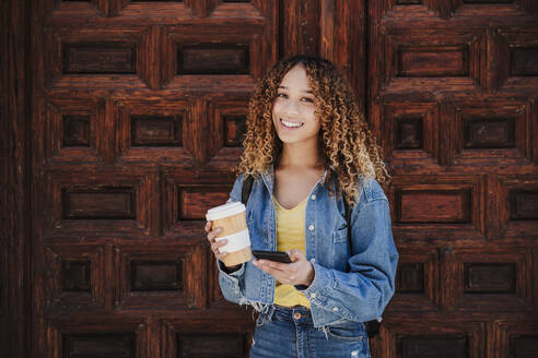 Junge Frau mit wiederverwendbarem Becher, die ihr Smartphone in der Hand hält, während sie vor einer Holztür steht - EBBF03670