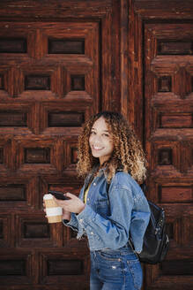 Lächelnde junge Frau in Jeansjacke an einer Holztür stehend - EBBF03671