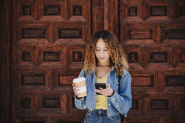 Curly haired woman holding reusable cup using smart phone by wooden door - EBBF03672