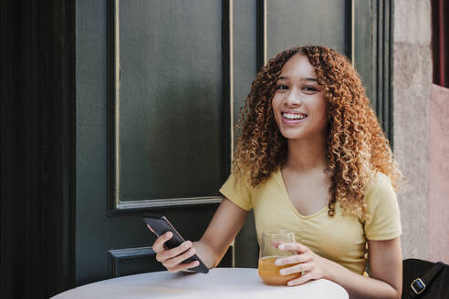 Lächelnde Frau mit Orangensaft, die ihr Smartphone hält, während sie in einem Straßencafé sitzt - EBBF03678