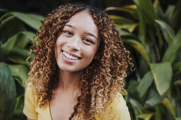 Smiling woman with curly hair near plants - EBBF03683