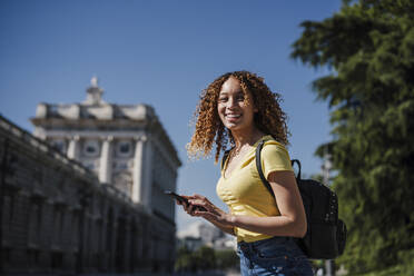 Smiling woman with backpack holding smart phone in city - EBBF03693