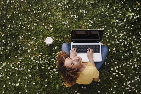 Young woman looking up while sitting with laptop on meadow amidst flowers - EBBF03699