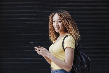 Young woman with backpack holding smart phone in front of shutter - EBBF03706