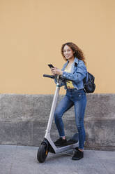 Young woman with electric push scooter holding smart phone while standing near wall - EBBF03708