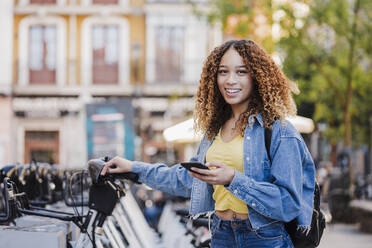 Smiling woman with smart phone standing near bicycle parking station - EBBF03723