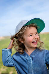 Happy blond girl in cap touching head and looking away against blue sky in summer in meadow - ADSF24545