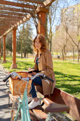 Young thoughtful female taking notes in planner on wooden bench near bicycle in park in daytime - ADSF24552