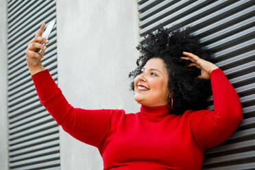 Side view of plump smiling female in bright wear making a self portrait while leaning on ribbed wall in town - ADSF24594