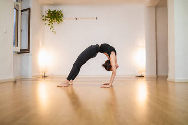 Seitenansicht einer nicht erkennbaren flexiblen Frau in Sportkleidung, die in Urdhva Dhanurasana-Pose steht, während sie Yoga auf glänzendem Boden übt - ADSF24615