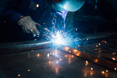 Crop gesichtslosen Arbeiter in Handschuhen und Uniform Schweißen Metallteile auf dem Tisch in der Nähe von Konstruktionen in der Fabrik - ADSF24626