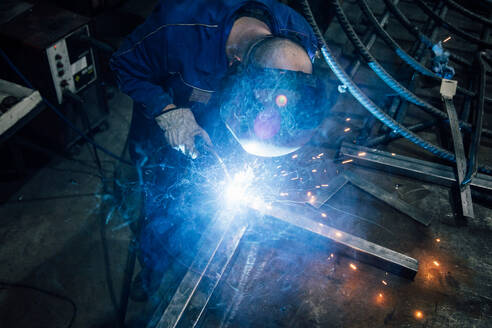 Crop gesichtslosen Arbeiter in Handschuhen und Uniform Schweißen Metallteile auf dem Tisch in der Nähe von Konstruktionen in der Fabrik - ADSF24631