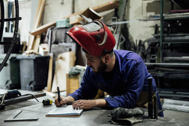 Seriöser bärtiger männlicher Arbeiter in Schutzhelm und Overall macht sich Notizen in einem Notizbuch mit Stift auf einem Tisch in der Nähe von Metallkonstruktionen in einer hellen Garage - ADSF24637