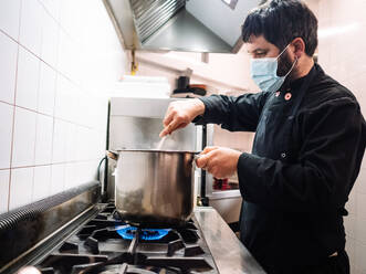 Seitenansicht eines professionellen männlichen Kochs mit Maske, der ein Gericht in einem Kochtopf umrührt, während er auf dem Herd in einer Restaurantküche kocht - ADSF24653