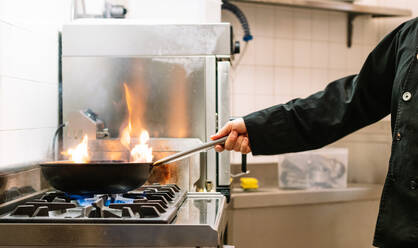 Side view of crop faceless cook preparing dish in frying pan on stove while working in kitchen of restaurant - ADSF24655